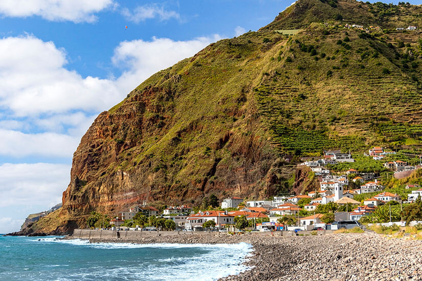 Eine kleine Küstensiedlung mit weißen Häusern und roten Dächern liegt zwischen Meer und einem steilen, grünen Berghang. Der Strand besteht aus dunklen Kieselsteinen, an denen die Wellen anrollen. Über den Häusern zieht sich eine Straße entlang des Hanges durch die terrassierte Landschaft. Madeira Madalena do Mar