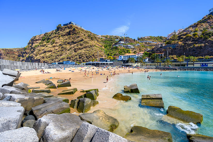 Ein Sandstrand mit vielen Badegästen liegt an einer geschützten Bucht mit klarem, türkisfarbenem Wasser. Große Betonblöcke und Felsen befinden sich im Vordergrund am Rand der Küste. Dahinter steigen steile Hügel mit Straßen, Häusern und Terrassenlandschaften an. Madeira Calheta Strand