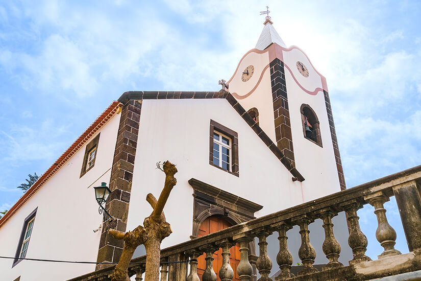 Eine weiße Kirche mit dunklen Steineinfassungen und einem kleinen Glockenturm steht vor einem bewölkten Himmel. Der Turm trägt eine Uhr und wird von einem Kreuz gekrönt. Im Vordergrund verläuft ein steinernes Geländer entlang einer Treppe oder Terrasse. Ponta do Sol Kirche