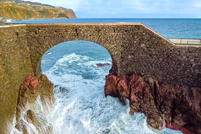 Eine steinerne Brücke mit einem großen Rundbogen überspannt eine felsige Küstenstelle. Unter dem Bogen schlagen Wellen gegen die Felsen und wirbeln weißes Meerwasser auf. Im Hintergrund sind das offene Meer und eine steile Küste mit Häusern zu erkennen. Cais da Ponta do Sol