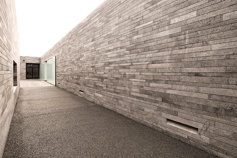 Ein langer, schmaler Gang verläuft zwischen hohen, grauen Steinwänden mit klarer, minimalistischer Architektur. Am Ende des Weges befindet sich ein moderner Eingangsbereich mit großen Glasflächen. Madeira Kunstmusem