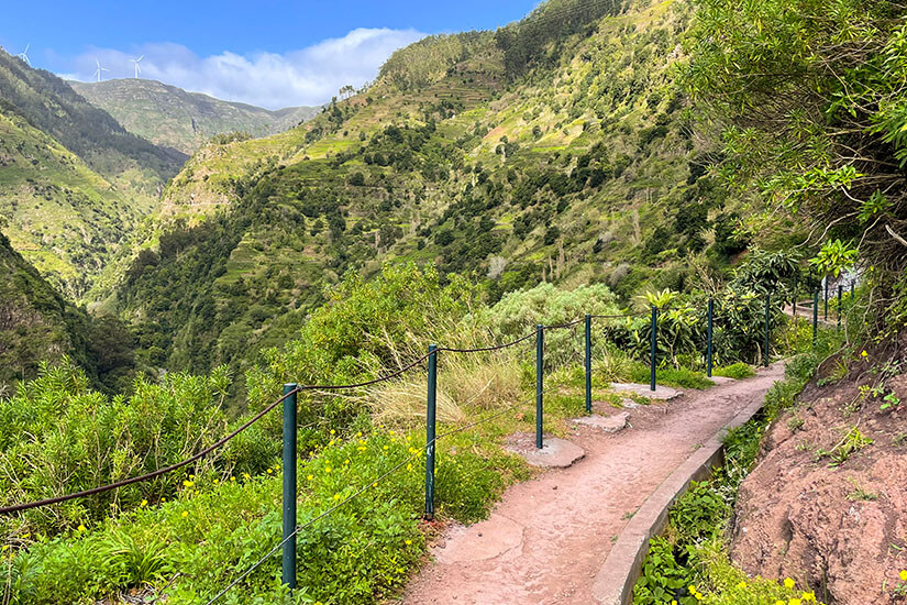 Ein schmaler Wanderweg führt entlang eines grünen Berghangs und ist mit einem Geländer gesichert. Die Landschaft besteht aus steilen Tälern, terrassierten Flächen und dichter Vegetation. In der Ferne sind auf einem Bergkamm mehrere Windräder zu erkennen. Ponta do Sol Levada Nova