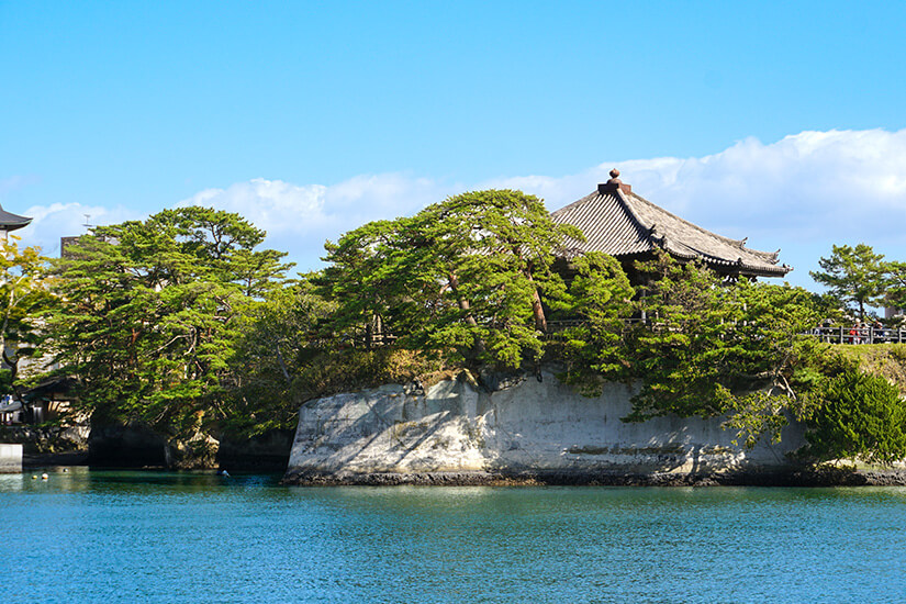 Ein traditioneller japanischer Tempel thront malerisch auf einer bewachsenen Felsinsel inmitten türkisfarbenen Wassers. Die markanten Kiefern und die weiße Felsklippe verleihen der Szene eine fast unwirkliche Schönheit. Matsushima bei Sendai zählt zu den „Drei schönsten Landschaften Japans. Sendai Japan Matsushima