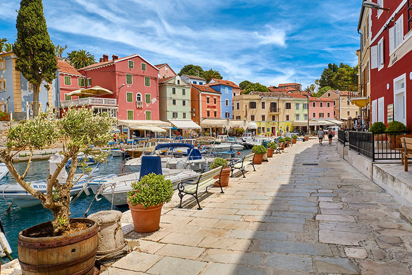 Eine Uferpromenade mit gepflastertem Weg verläuft entlang eines kleinen Hafens mit Booten. Bunte Häuser in Pastelltönen säumen die Straße und verleihen der Szene ein mediterranes Flair. Pflanzen in Töpfen, Sitzbänke und einige Spaziergänger beleben die Umgebung. Veli Lošinj Promenade