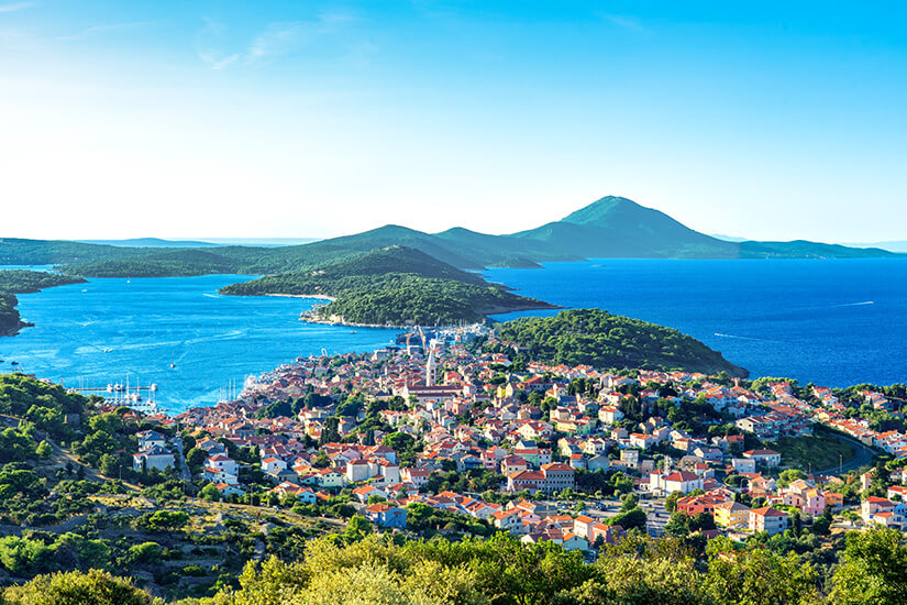 Eine Küstenstadt mit vielen dicht stehenden Häusern breitet sich auf einer Halbinsel aus. Umgeben ist sie von tiefblauem Meer und mehreren grünen Inseln. Im Hintergrund ragt eine bergige Landschaft unter klarem Himmel auf. Lošinj Insel