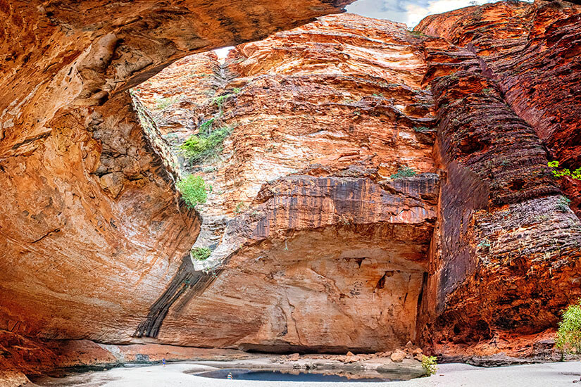 Eine gewaltige, überhängende Felswand aus rot-orange gebändertem Sandstein bildet eine natürliche Kathedrale – typisch für den Purnululu Nationalpark in Westaustralien. Die beeindruckenden Formationen der Bungle Bungles gehören zum UNESCO-Welterbe und zählen zu den einzigartigen Naturwundern des Kontinents. Dieser australische Nationalpark ist ein absolutes Highlight für jeden Besucher Westaustraliens.