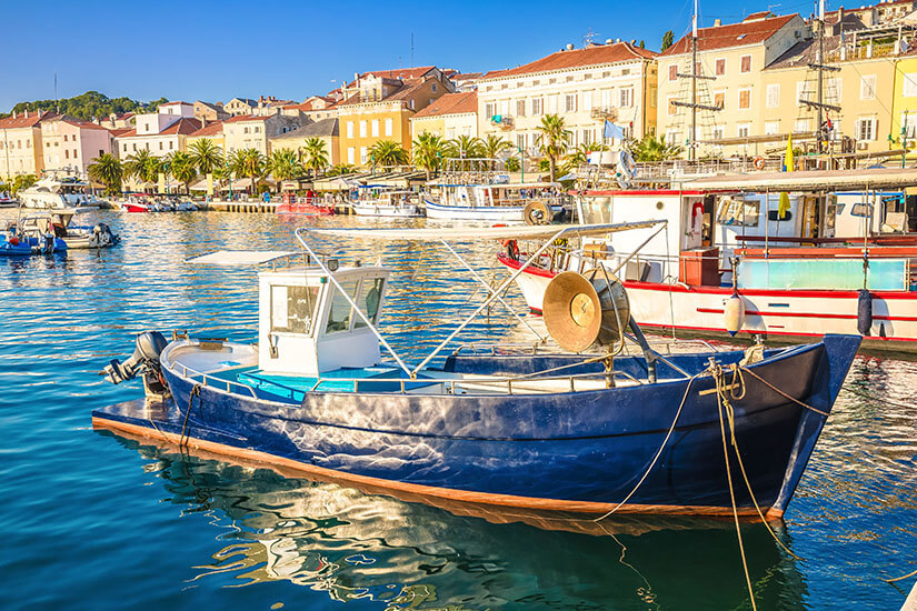 Ein kleines Boot liegt im ruhigen Hafenwasser und ist mit Seilen am Ufer befestigt. Dahinter befinden sich weitere Boote sowie eine Reihe von Häusern mit hellen Fassaden und roten Dächern. Palmen entlang der Promenade verstärken die mediterrane Atmosphäre. Kroatien Mali Losinj
