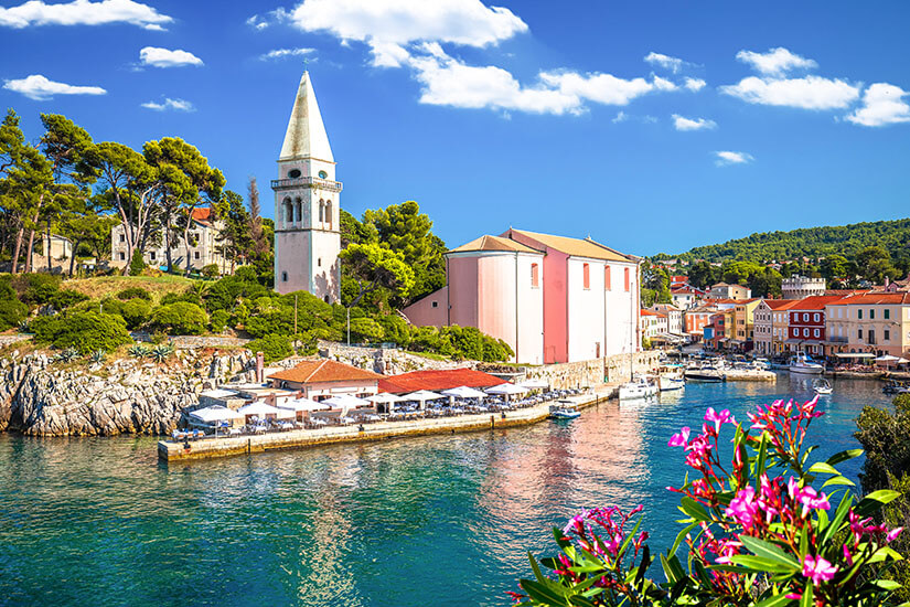 Ein malerischer Küstenort mit Hafen liegt am klaren Wasser, umgeben von bunten Häusern und viel Grün. Eine Kirche mit hohem Turm ragt über die Gebäude hinaus. Im Vordergrund sorgen blühende Pflanzen für zusätzliche Farbakzente. Veli Losinj