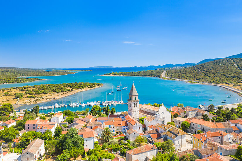 Eine kleine Stadt mit roten Ziegeldächern liegt an einer schmalen Meerenge mit türkisfarbenem Wasser. Im Hafen sind zahlreiche Segelboote und Yachten zu sehen. Hügelige, grüne Landschaften umgeben die Küste unter einem klaren Himmel. Insel Lošinj Osor