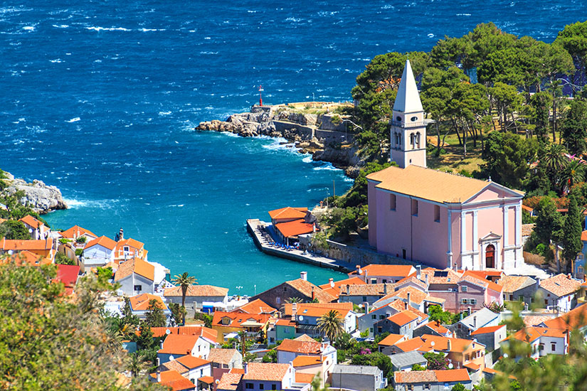 Ein Küstenort mit dicht stehenden Häusern und roten Dächern liegt an einer kleinen Bucht mit tiefblauem Wasser. Eine große Kirche mit Turm steht nahe am Ufer und prägt das Ortsbild. Felsen, Bäume und ein kleiner Hafenbereich rahmen die Szenerie ein. Veli Losinj Kirche