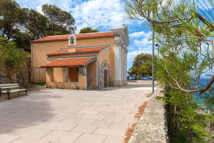 Eine kleine Kapelle mit heller Fassade und roten Ziegeldächern steht auf einer gepflasterten Fläche nahe der Küste. Bäume und Sträucher umgeben das Gebäude, während das Meer im Hintergrund sichtbar ist. Lošinj Kirche Annunziata