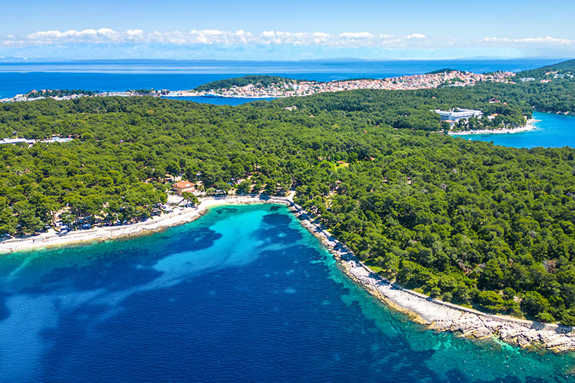 Eine türkisfarbene Bucht wird von dicht bewaldeten Küstenstreifen umgeben. Entlang des Ufers verlaufen helle Strände und kleine Wege durch die Natur. In der Ferne erstreckt sich eine weitere Siedlung auf einer schmalen Landzunge ins Meer hinaus. Cikat Bucht Mali Losinj