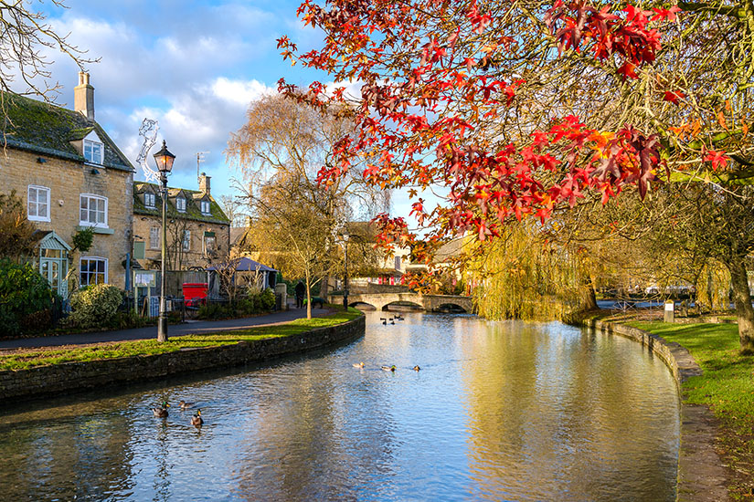 Malerische herbstliche Ansicht des Flusses Windrush in Bourton-on-the-Water, Cotswolds, mit Enten auf dem Wasser, historischen Steinhäusern und einer Bogenbrücke unter blauem Himmel. Cotwolds England Bourton on the Water