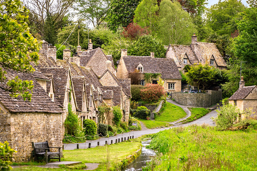 Berühmte historische Steinhäuser der Arlington Row in Bibury, Cotswolds, mit charakteristischen spitzen Giebeln, üppigem Grün und einem kleinen Wasserlauf im Vordergrund. Cotswolds England Bibury