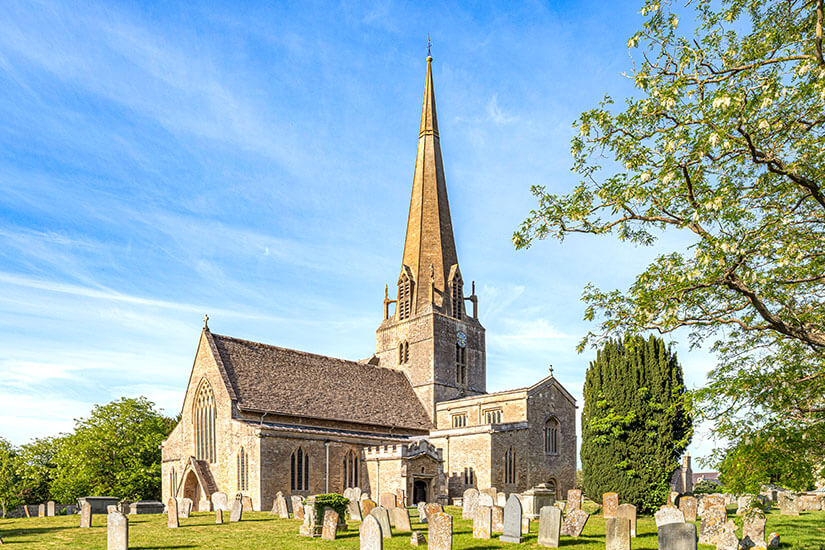 Historische St Mary's Church in Bampton, Cotswolds, mit hohem Kirchturm und dem umliegenden, grasbewachsenen Friedhof mit alten Grabsteinen bei strahlend blauem Himmel. England St Marys Church Bampton Cotswolds England St Marys Church Bampton