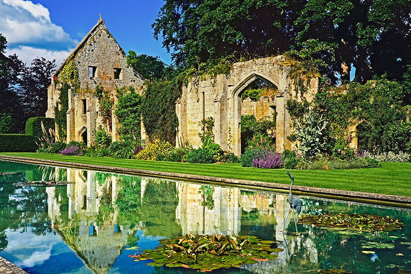 Atemberaubende Ansicht der historischen, efeubewachsenen Scheunenruine von Sudeley Castle, die sich perfekt in einem langen Spiegelteich mit Seerosen und einer bronzenen Reiherstatue spiegelt. Ein gepflegter Garten mit bunten Blumen und grünen Hecken umrahmt die Szene unter leicht bewölktem Himmel. Cotswolds England Scheine Sudeley Castle