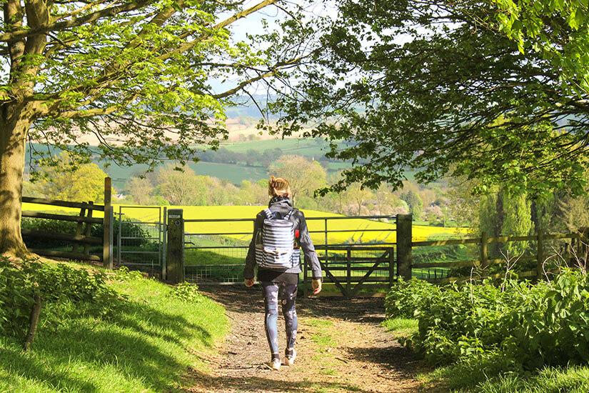 Rückansicht einer Wanderin mit Rucksack, die auf einem Feldweg durch ein Holztor geht. Im Hintergrund erstreckt sich die Hügellandschaft der Cotswolds mit einem leuchtend gelben Rapsfeld unter Bäumen. Cotwolds England Wandern