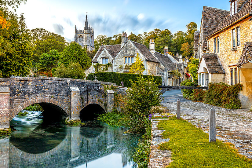 Blick auf die historische Steinbrücke über den Fluss Bybrook in Castle Combe mit Spiegelungen im Wasser, den typischen honigfarbenen Steinhäusern und dem Kirchturm von St. Andrew's im Hintergrund. Cotswolds England Castle Combe