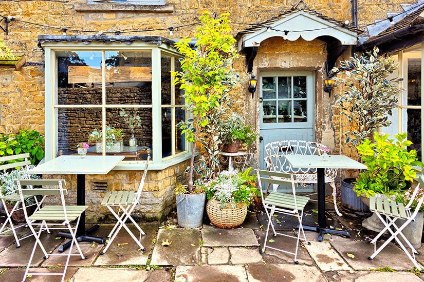 Außenansicht eines traditionellen, honigfarbenen Steinhauses in den Cotswolds mit einem gemütlichen Café-Eingang. Davor stehen hellgrüne Metalltische und -stühle auf einem Steinpflaster, umgeben von Topfpflanzen und Lichterketten. Cotswolds England Teestube
