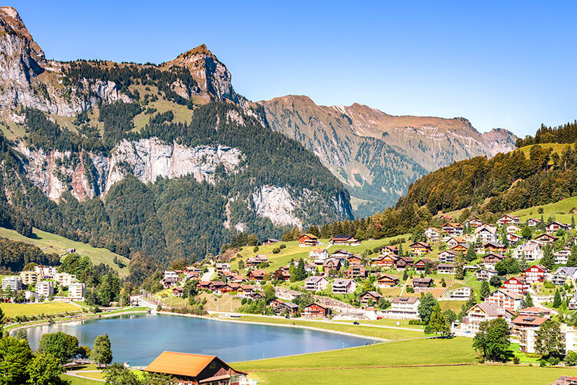 Das Bergdorf Engeldorf in der Schweiz ist zu sehen, mit hellblauem See, Häusern auf grünen Wiesen und gewaltigem Bergmassiv im Hintergrund. Engelberg Schweiz Willkommen