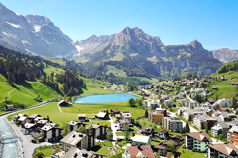 Das idyllische Dorf Engelberg umgeben von Gipfeln ist zu sehen. Grüne Almwiesen leuchten und in der Mitte glitzert ein See. Engelberg Schweiz Wetter