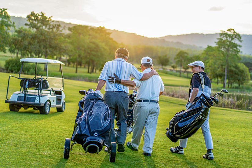 Drei Golfspieler spazieren im warmen Abendlicht eines Sonnenuntergangs über ein weitläufiges Fairway, zwei von ihnen mit Trolleys, einer mit einer Schultertasche, während ein Golfcart am Rand steht. Packliste Golfurlaub Gruppe