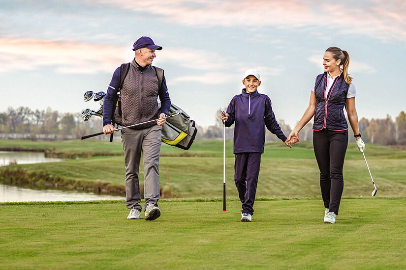 Vater, Mutter und Kind spazieren fröhlich über einen gepflegten Golfplatz. Alle drei in passenden dunkelblauen Outfits und mit Ausrüstung. Packliste Golfurlaub Familie