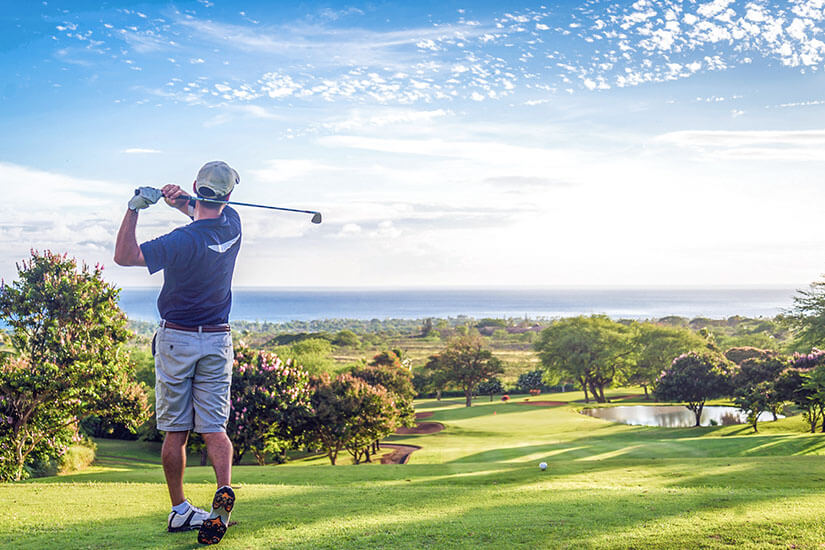 Ein Golfspieler führt einen eleganten Abschlag auf einem malerischen Golfplatz aus, der mit seinem satten Grün, blühenden Sträuchern und einem kleinen Teich eine atemberaubende Kulisse bietet. Packliste Golfurlaub Abschlag