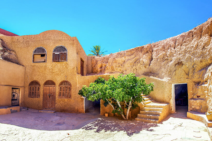 Ein traditionelles Lehmhaus mit abgerundeten Formen und kleinen Fenstern steht in einem Innenhof, der teilweise in den Fels gebaut ist. Eine Treppe führt zu einer höher gelegenen Ebene, während ein Baum in der Mitte etwas Schatten spendet. Matmata Hoehlendorf