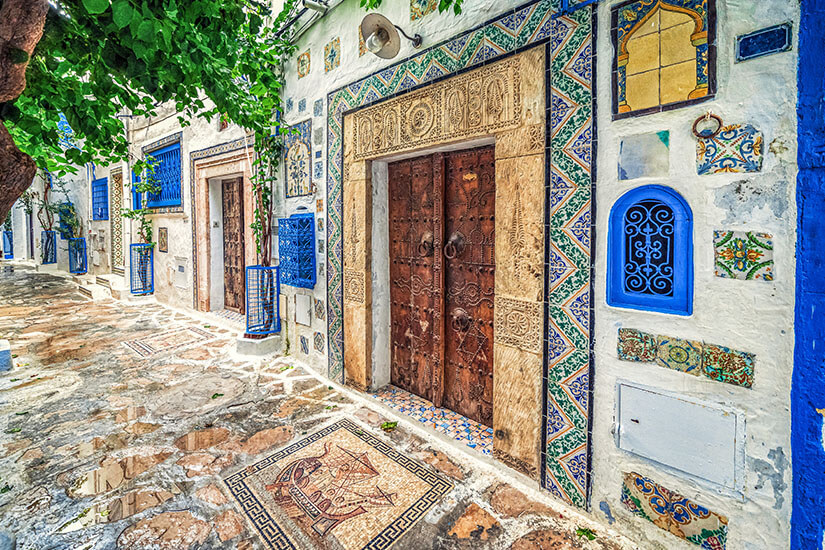 Verzierte Hausfassaden mit kunstvollen Fliesen und geschnitzten Holztüren prägen eine enge Gasse. Blaue Fensterrahmen und Ornamente setzen farbige Akzente auf den hellen Wänden. Der gepflasterte Boden ist ebenfalls dekorativ gestaltet und teilweise mit Mustern versehen. Tunesien Hammamet