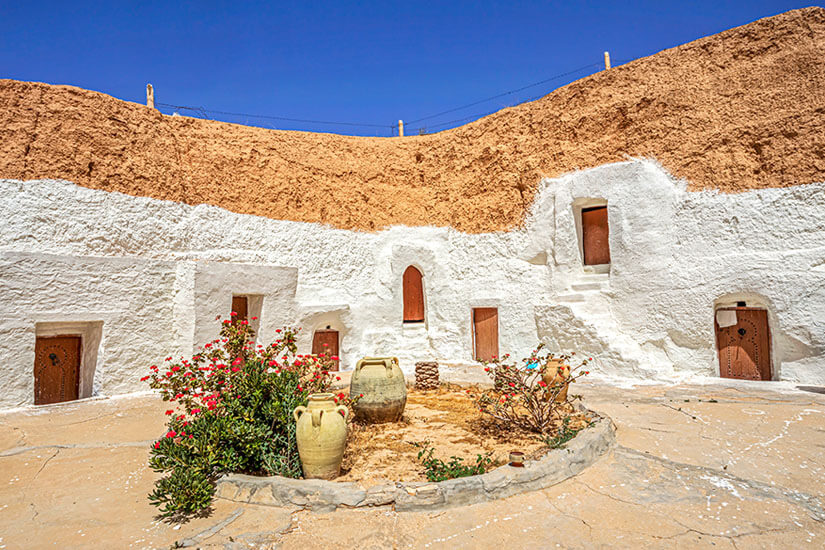 Ein runder Innenhof ist von in den Fels gehauenen Wohnräumen mit weißen Wänden umgeben. Mehrere Türen und kleine Fenster führen in die Höhlenräume. In der Mitte stehen Pflanzen und große Tonkrüge als Dekoration. Matmata Hoehlenwohnungen