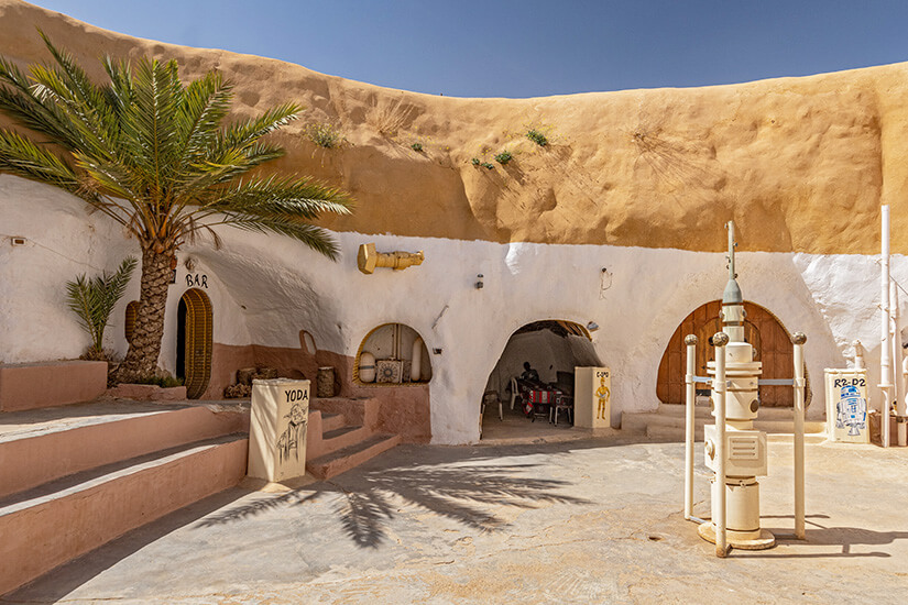 Ein sandfarbener Innenhof mit in den Fels gehauenen, weiß verputzten Räumen erinnert an eine Wüstenkulisse. Mehrere Türen und Bögen führen in die Höhlen, daneben stehen Requisiten und Figuren aus der „Star Wars“-Welt wie R2-D2 und Yoda. Eine Palme und einfache Sitzbereiche ergänzen die Szenerie. Matmata Star Wars