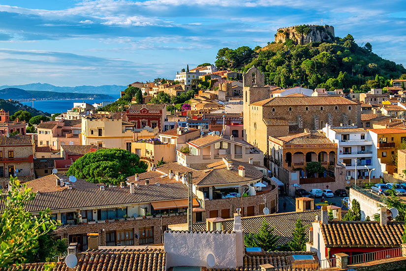 Die Altstadt von Begur ist zu sehen. Auf dem grünen Hügel thront die Burg. Häuser mit heller Fassade sind zu sehen. Im Hintergrund glitzert das Mittelmeer. Begur Spanien Willkommen