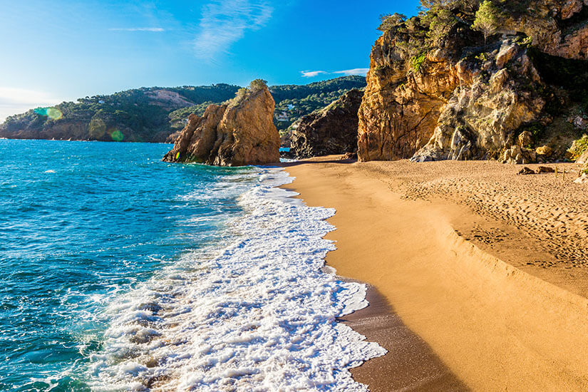 Ein goldener Sandstrand mit Felsen im Hintergrund. Das Wasser leuchtet blau. Begur Spanien Roja
