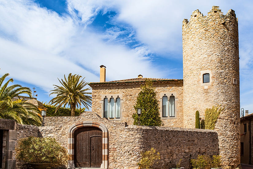 Ein ehemaliger Wehrturm aus hellem Stein thront neben einem Haus und einer Mauer mit Holztor. Begur Spanien Torre