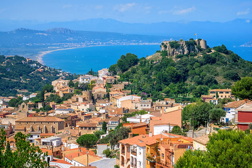 Ein Blick auf die Stadt Begur mit ihren roten Dächern und dem Castillo auf dem grünen Hügel. Im Hintergrund ist eine Bucht am Mittelmeer zu sehen. Begur Spanien Wetter