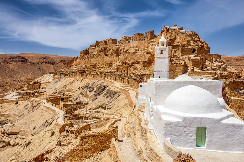 Ein Dorf aus Lehm- und Steinbauten zieht sich terrassenförmig einen kargen Hügel in der Wüste hinauf. Im Vordergrund steht ein kleines weißes Gebäude mit Kuppel und Minarett. Schmale Wege verbinden die Häuser und schlängeln sich durch die trockene Landschaft. Tunesien Chenini