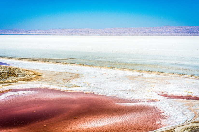 Eine weite, fast weiße Salzlandschaft erstreckt sich bis zum Horizont, unterbrochen von flachen Wasserflächen. Im Vordergrund hebt sich ein rötlich gefärbtes Becken deutlich vom hellen Salz ab. In der Ferne sind niedrige Berge unter einem klaren blauen Himmel zu erkennen. Matmata Salzsee