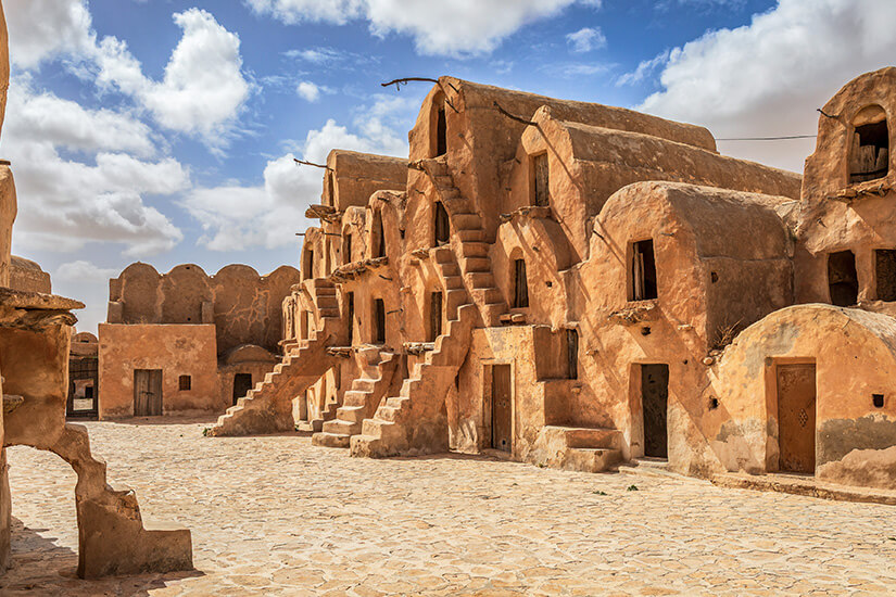 Lehmfarbene, mehrstöckige Gebäude mit gewölbten Formen und Außentreppen umgeben einen offenen Innenhof. Kleine Türen führen in die einzelnen Kammern, die wie Speicher oder Wohnräume wirken. Matmata Ksar Ouled Soltane