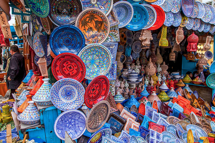 Ein Marktstand ist dicht gefüllt mit bunt bemalter Keramik, darunter Teller, Schalen und Tajine-Gefäße mit aufwendigen Mustern. Die leuchtenden Farben wie Blau, Rot und Weiß dominieren das Bild und erzeugen ein lebendiges Gesamtbild. Matmata Berber