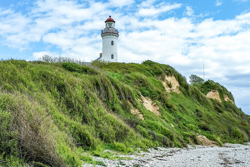 Blick von einem Kiesstrand hinauf zu einer steilen, grasbewachsenen Klippe, auf deren Gipfel der weiße Leuchtturm Vesborg Fyr mit seiner roten Laterne steht, unter einem teilweise bewölkten blauen Himmel. Samsoe Daenemark Vesborg Fyr