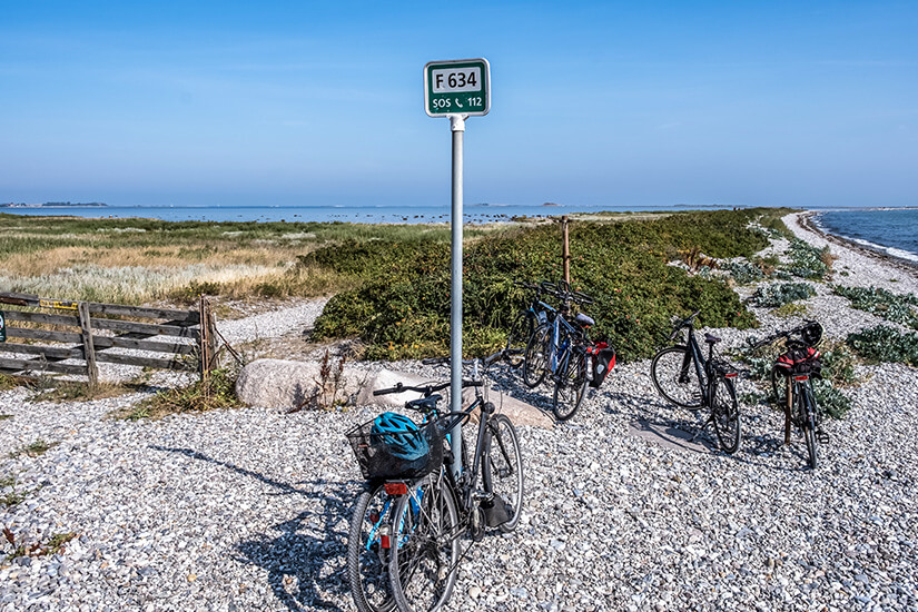 Mehrere Fahrräder parken an einem Metallpfosten mit einem SOS-Schild auf einem hellen Kiesweg direkt am Meer. Im Hintergrund erstreckt sich eine schmale Landzunge mit grüner Vegetation unter einem klaren blauen Himmel. Samsoe Daenemark Radfahren