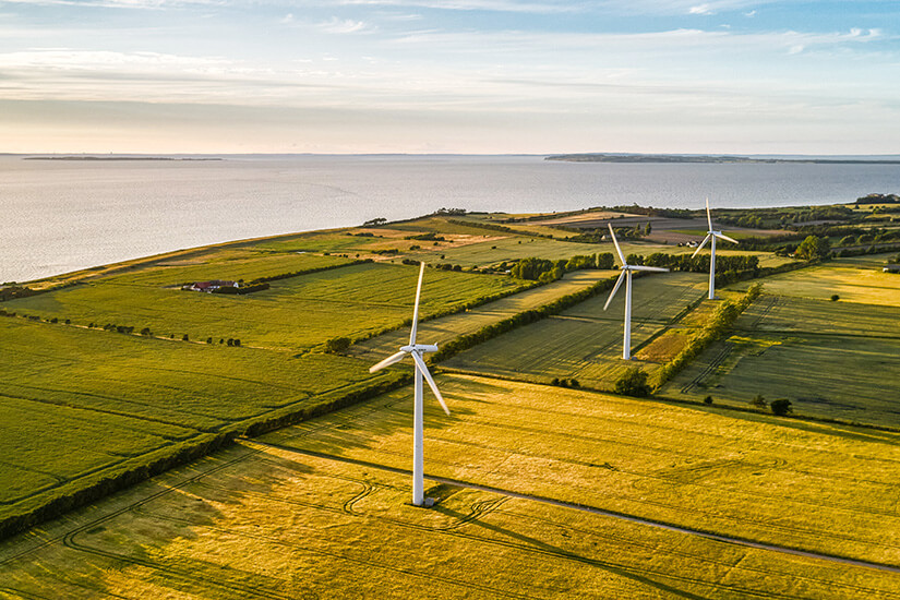 Eine Luftaufnahme von drei großen, weißen Windrädern, die in einer Reihe auf weiten, goldgelben und grünen Feldern stehen. Die Felder erstrecken sich bis zur Küste des ruhigen Meeres unter einem blauen Himmel mit weichen Wolken, aufgenommen im sanften Licht der Abenddämmerung. Samsoe Daenemark Windpark