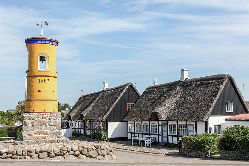 Glockenturm in Nordby, Samsø, aus dem Jahr 1857 auf Steinfundament, dahinter reetgedeckte Fachwerkhäuser unter blauem Himmel. Daenemark Nordby Glockenturm Samsoe Daenemark Nordby Glockenturm