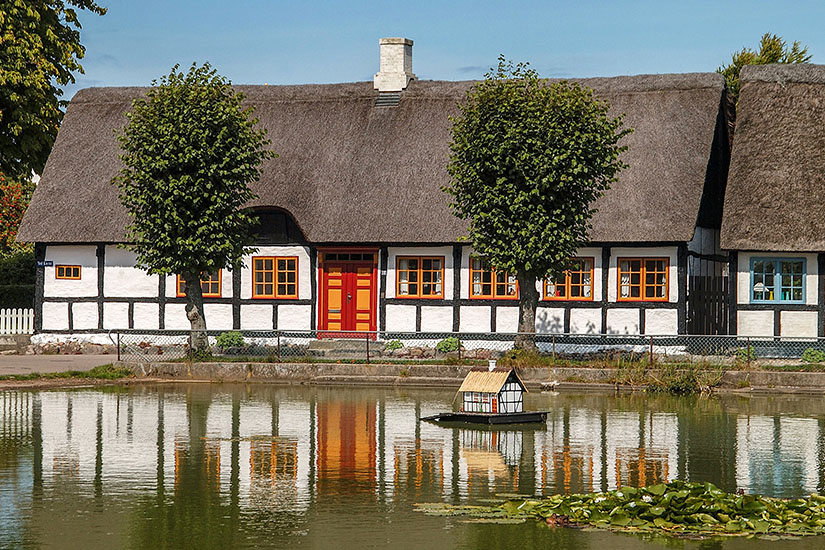 Ein idyllischer Dorfteich in Nordby auf Samsö, in dem sich ein weißes Fachwerkhaus mit Reetdach spiegelt. Auf dem Wasser schwimmt ein kleines Modellhaus im gleichen Stil, umgeben von Seerosen. Daenemark Nordby Teich Samsoe Daenemark Nordby Teich