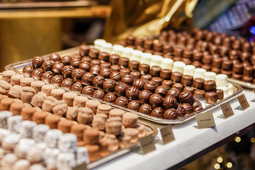 Reihen von fein arrangierten Pralinen und Trüffeln liegen auf Tabletts in einer Auslage. Verschiedene Sorten aus heller und dunkler Schokolade sind sorgfältig geformt und dekoriert. Kleine Beschriftungen weisen auf die jeweiligen Varianten hin. Schweiz Souvenir Schokolade