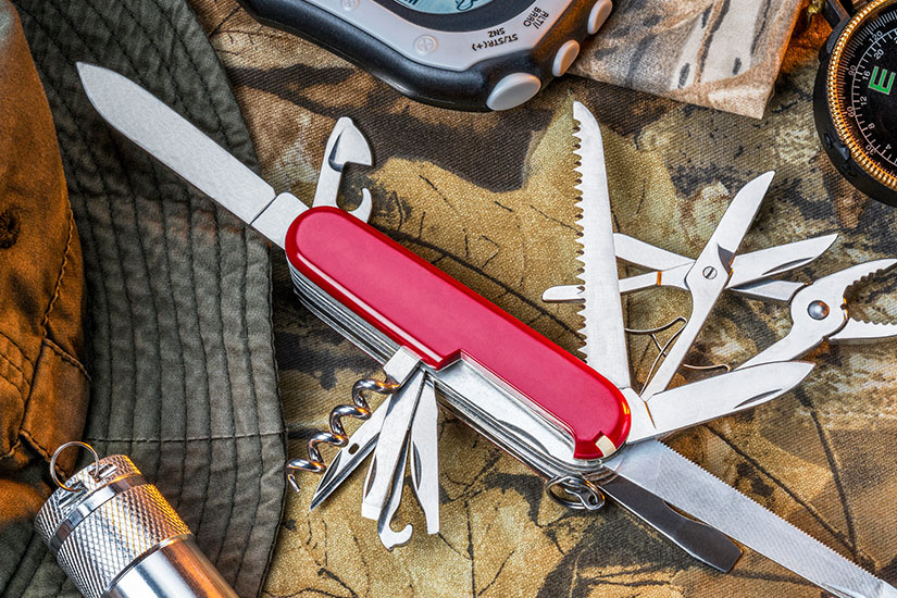 Ein rotes Multifunktions-Taschenmesser liegt aufgeklappt und zeigt verschiedene Werkzeuge wie Messer, Schere und Säge. Es liegt auf einer Karte neben Outdoor-Gegenständen wie Kompass und Taschenlampe. Schweiz Souvenirs Taschenmesser