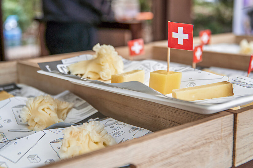 Kleine Stücke und Späne von Käse liegen auf Tabletts, einige mit kleinen Fähnchen mit Schweizer Kreuz dekoriert. Messer liegen bereit, sodass man die Proben direkt schneiden oder probieren kann. Schweiz Mitbringsel Käse