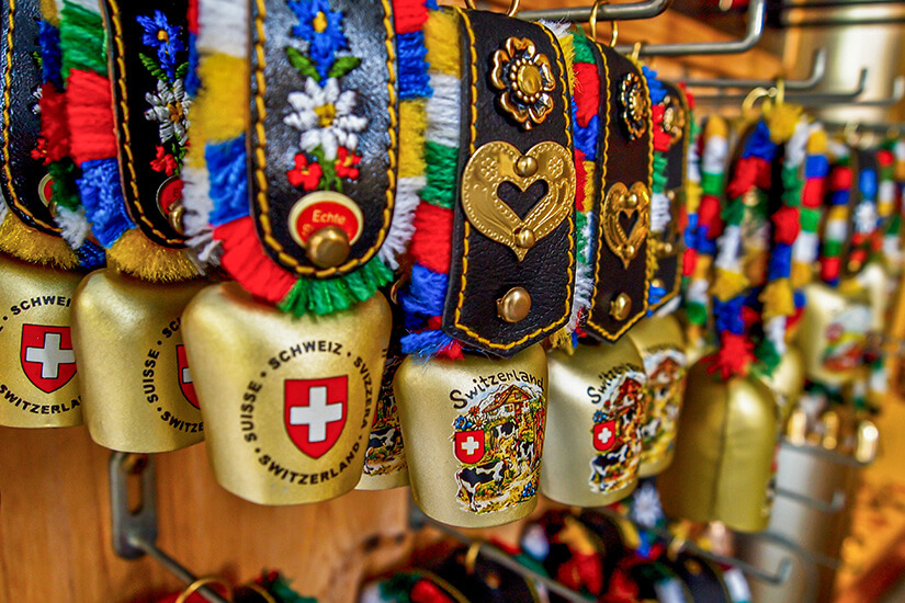 Bunte Kuhglocken mit verzierten Lederbändern hängen dicht nebeneinander an einem Verkaufsstand. Die Bänder sind mit Stickereien, Mustern und kleinen Metallornamenten geschmückt. Auf den Glocken sind Schweizer Symbole und Schriftzüge zu sehen. Souvenirs Kuhglocken