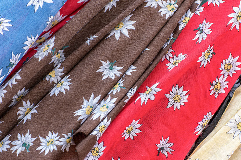 Mehrere Stoffe oder Tücher liegen nebeneinander, bedruckt mit kleinen Edelweißblüten. Die Farben variieren zwischen Rot, Blau, Braun und Beige. Mitbringsel Edelweiß